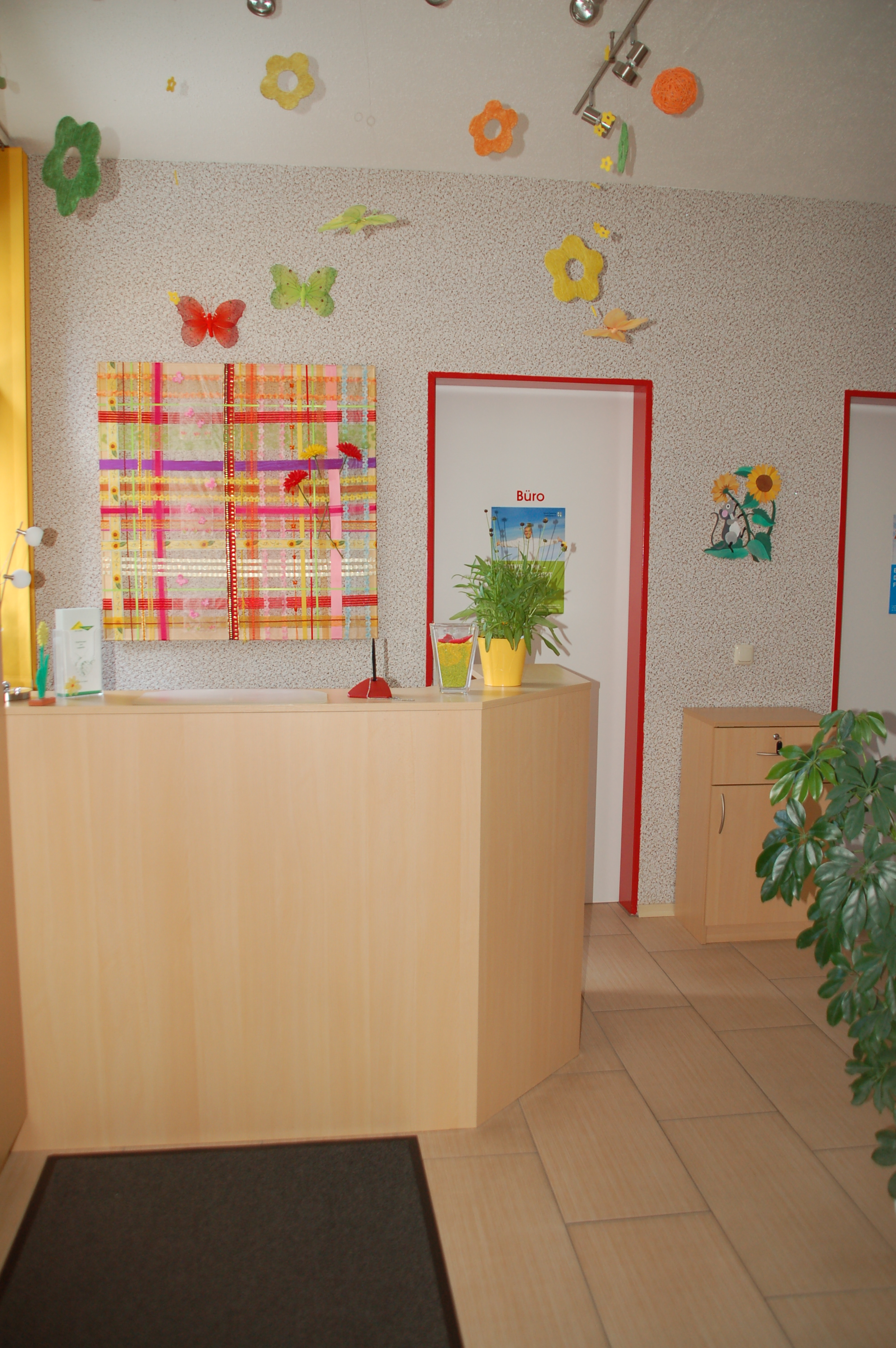 Empfangstheke in einem bunten Wartezimmer mit Blumentöpfen, dekorativen Schmetterlingen und Blumen an der Wand und Tür mit Schild 'Büro'.