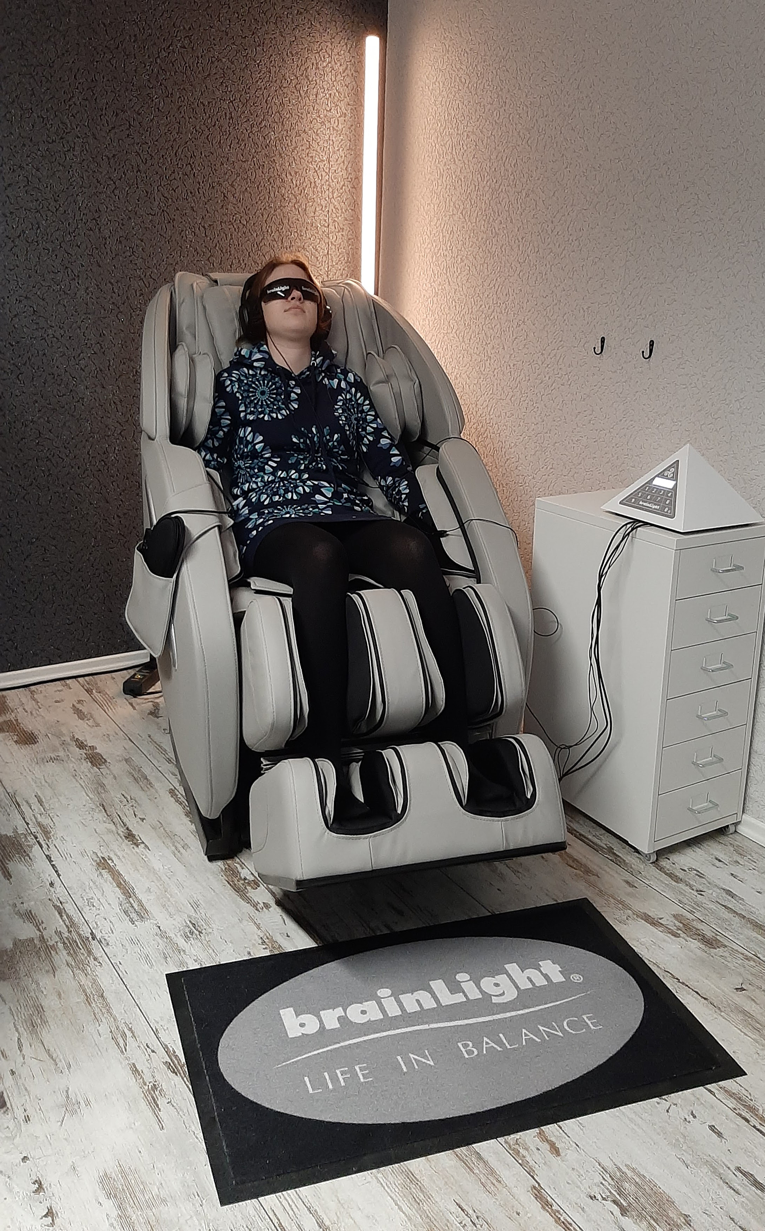 Eine Person sitzt entspannt in einem grauen Massage- und Wellnessstuhl, trägt eine Augenmaske und Kopfhörer, neben dem Stuhl steht ein schrankähnliches Gerät mit einem pyramidenförmigen Display, darunter liegt eine Matte mit der Aufschrift ‚brainLight LIFE IN BALANCE‘.