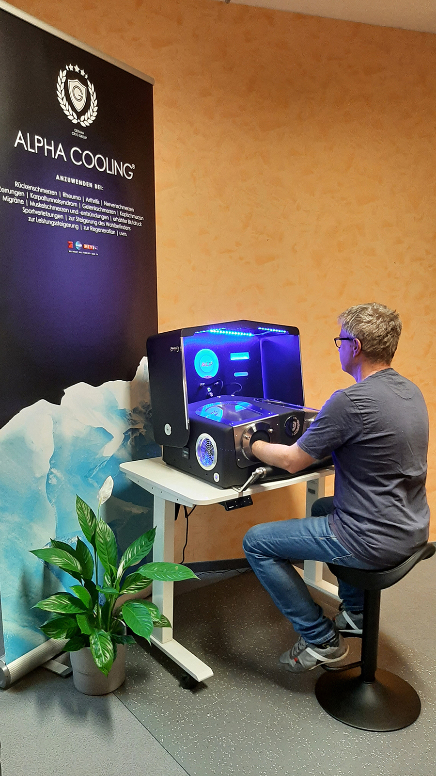Ein Mann sitzt auf einem Hocker und steckt seine Hände in ein Gerät mit blauer Beleuchtung auf einem Tisch neben einem Banner für Alpha Cooling.
