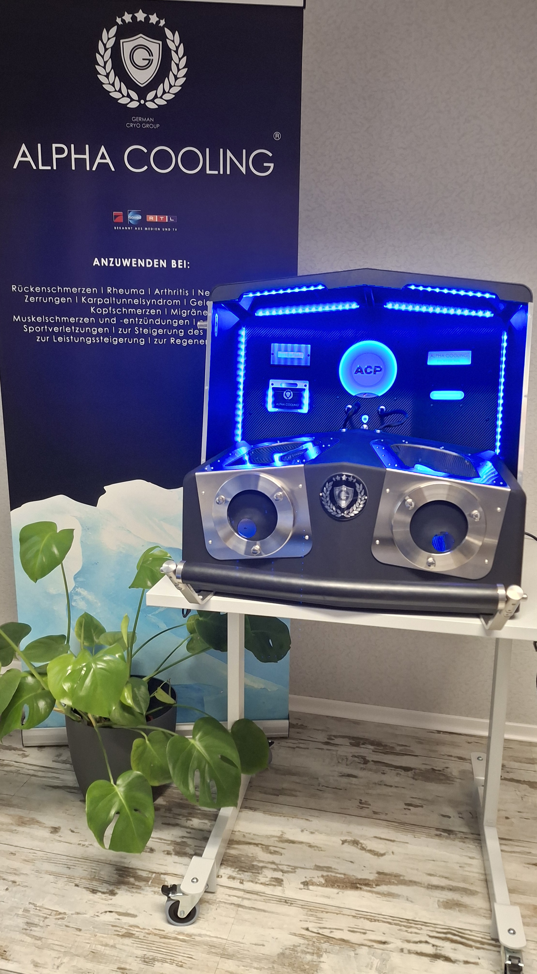 Alpha Cooling Gerät mit blauer LED-Beleuchtung auf einem weißen Ständer neben einem Banner und einer grünen Pflanze.