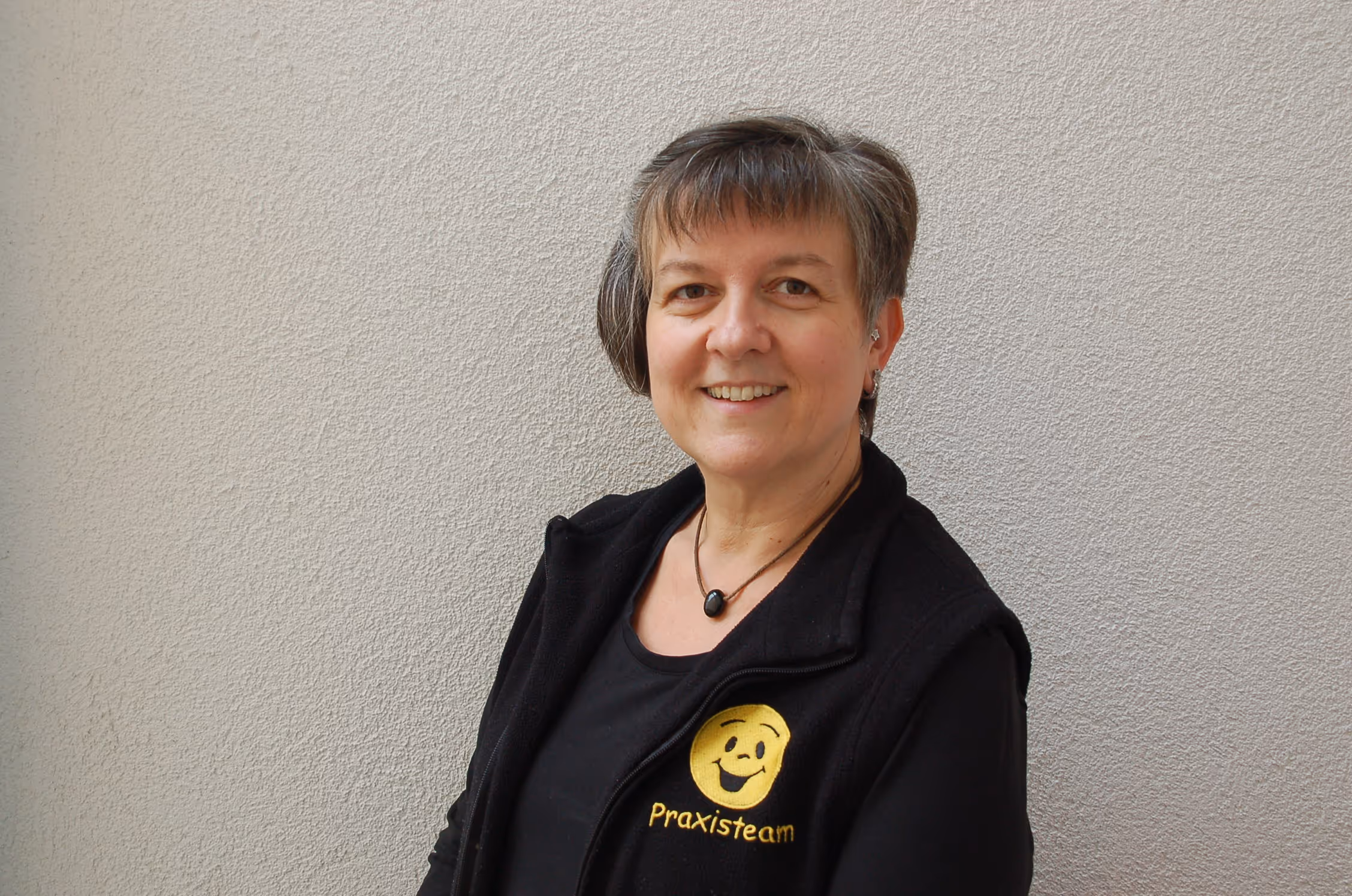 Lächelnde Frau mit kurzem Haar trägt eine schwarze Weste mit gelbem Praxisteam-Logo vor einer hellen Wand.