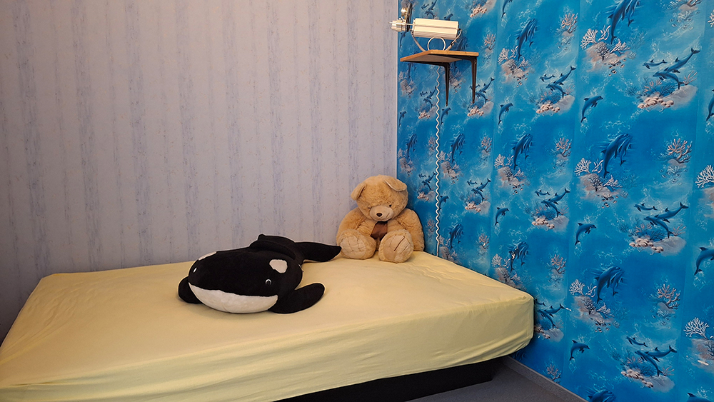Ein einfaches Bett mit gelbem Laken, einem Plüsch-Orca und einem braunen Teddybär vor einer blauen Wand mit Delfin- und Korallenmuster.