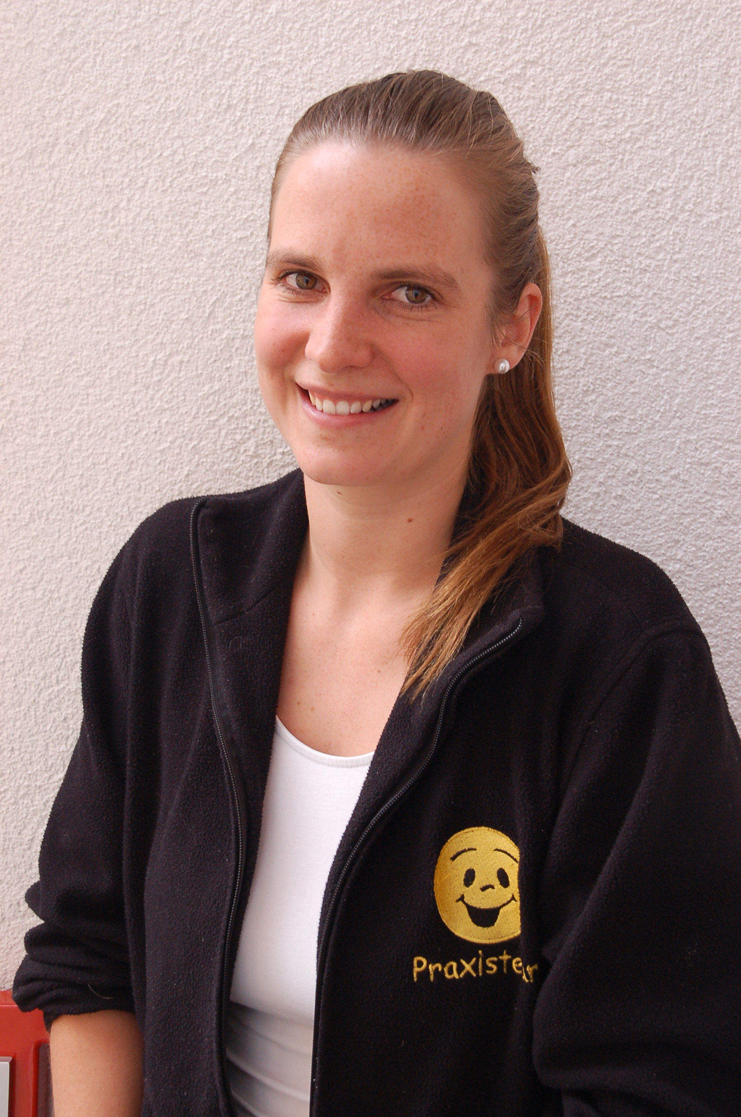 Junge Frau mit zusammengebundenem Haar und einem schwarzen Pullover mit gelbem Smiley-Logo und dem Schriftzug 'Praxisteam' lächelt vor einer hellen Wand.