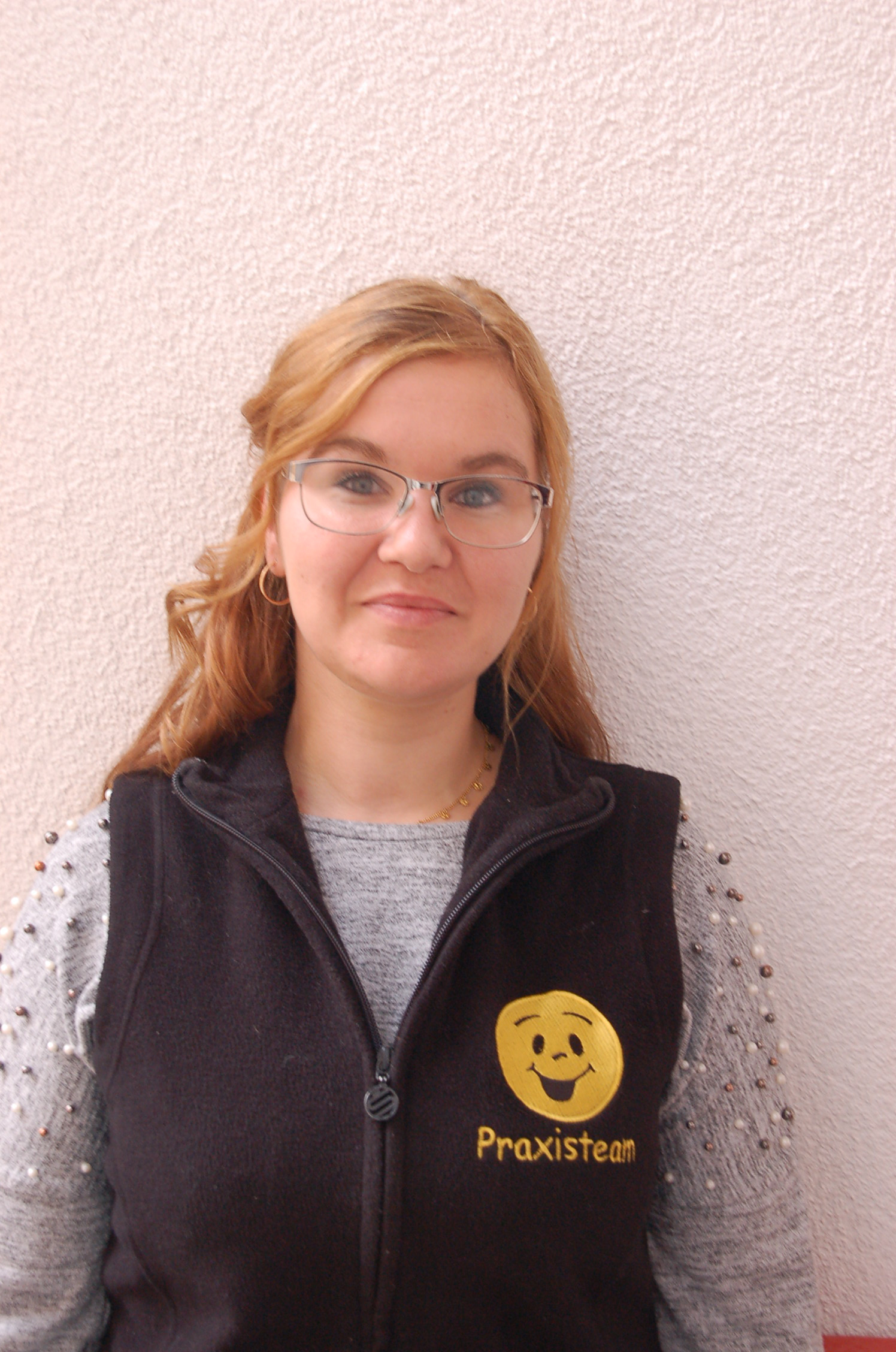 Frau mit hellbraunen Haaren und Brille trägt eine schwarze Weste mit gelbem Smiley-Logo und der Aufschrift ‚Praxisteam‘.