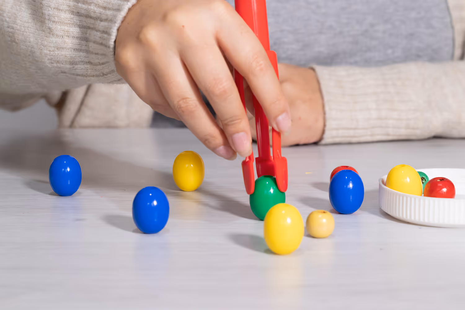Hand hält rote Zange und hebt grünes Kunststoffei von Tisch mit bunten Plastikfingern.
