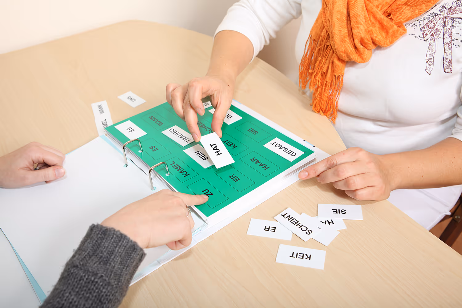 Zwei Personen sortieren beschriftete Karten mit deutschen Wörtern auf einem farbigen Blatt im Ordner.