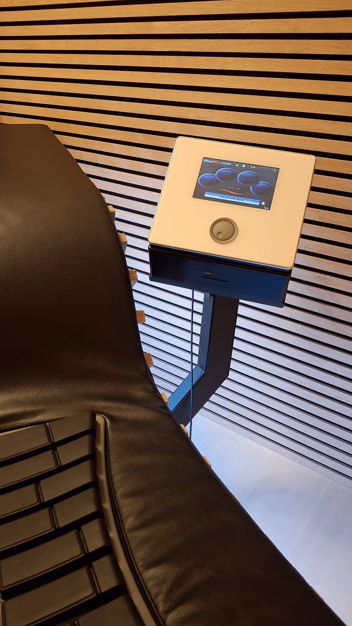 Schwarze Liege mit Steuergerät in einem Raum mit Holzlamellenwand.