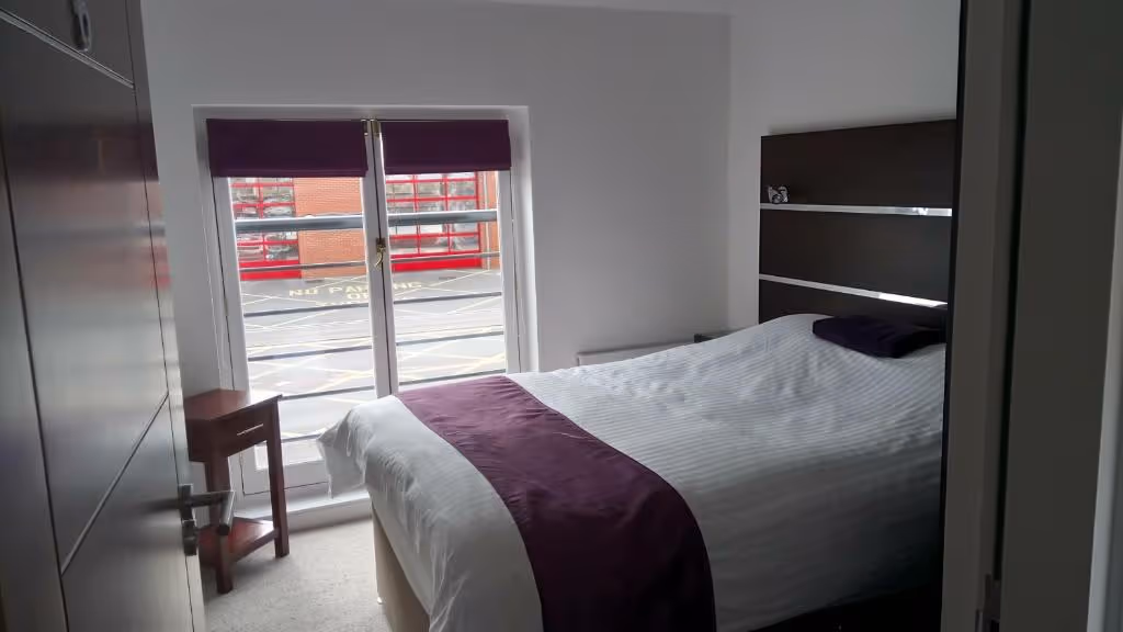 A bright and modern guest room at Rooms At The Inn. The room is furnished with a comfortable bed with purple accents, contemporary dark wood furniture including a wardrobe and headboard, and a large window for natural light.