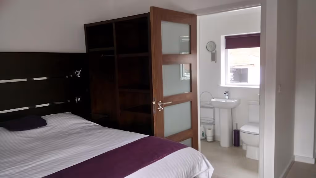 A contemporary guest room at Rooms At The Inn, highlighting the efficient layout with dark wood furnishings, an open wardrobe for storage, and a bright, adjacent ensuite bathroom with modern fixtures.