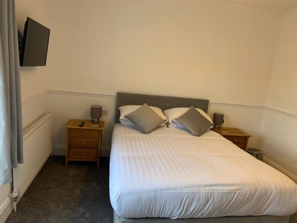 A neatly made double bed with white linens and two grey accent pillows, flanked by wooden nightstands with matching grey lamps in a cozy hotel room at Room At The Inn, Retford. A wall-mounted TV is visible on the left above a radiator, and the room features neutral tones with white walls and grey carpeting.