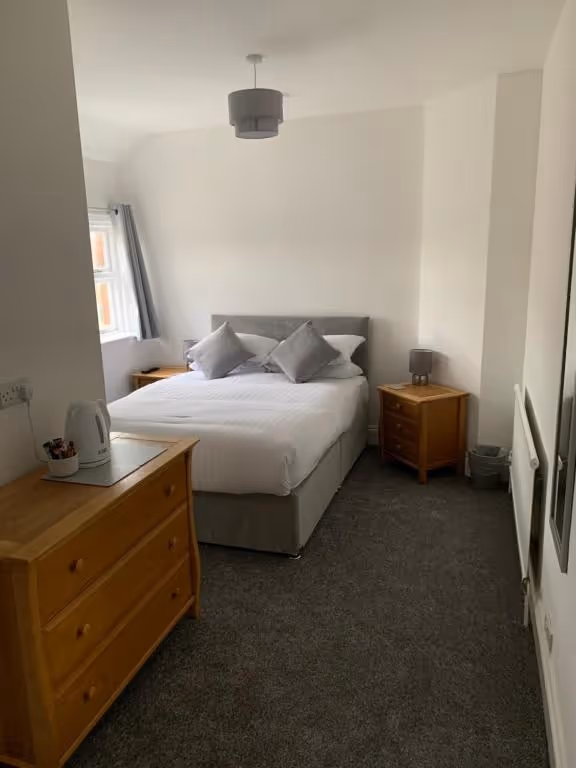 A neatly made double bed with white linens and two grey accent pillows, flanked by wooden nightstands with matching grey lamps in a cozy hotel room at Room At The Inn, Retford. A wall-mounted TV is visible on the left above a radiator, and the room features neutral tones with white walls and grey carpeting.