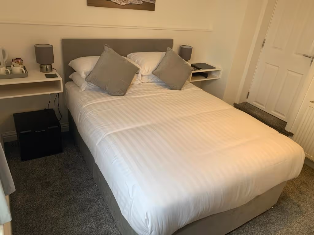 A cozy hotel room at Room At The Inn, Retford, featuring a neatly made double bed with white striped linens and two grey accent pillows. Modern floating bedside shelves hold matching grey lamps, a tea and coffee tray, and a tablet or book. A small black mini fridge is beneath one shelf, and a white door and carpeted step are visible in the background.