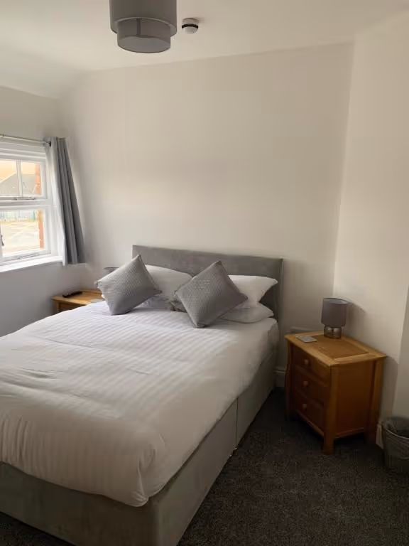 A neatly made double bed with white linens and two grey accent pillows, flanked by wooden nightstands with matching grey lamps in a cozy hotel room at Room At The Inn, Retford. A wall-mounted TV is visible on the left above a radiator, and the room features neutral tones with white walls and grey carpeting.