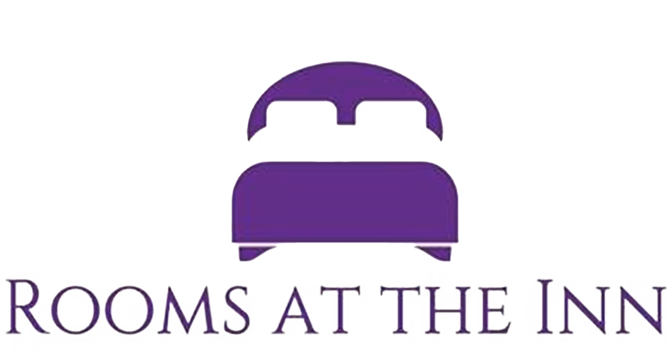 Logo for the accommodation "Rooms At The Inn". The logo consists of a stylized purple graphic of a double bed with a rounded headboard, positioned above the business name which is written in a purple, capitalised, serif typeface.