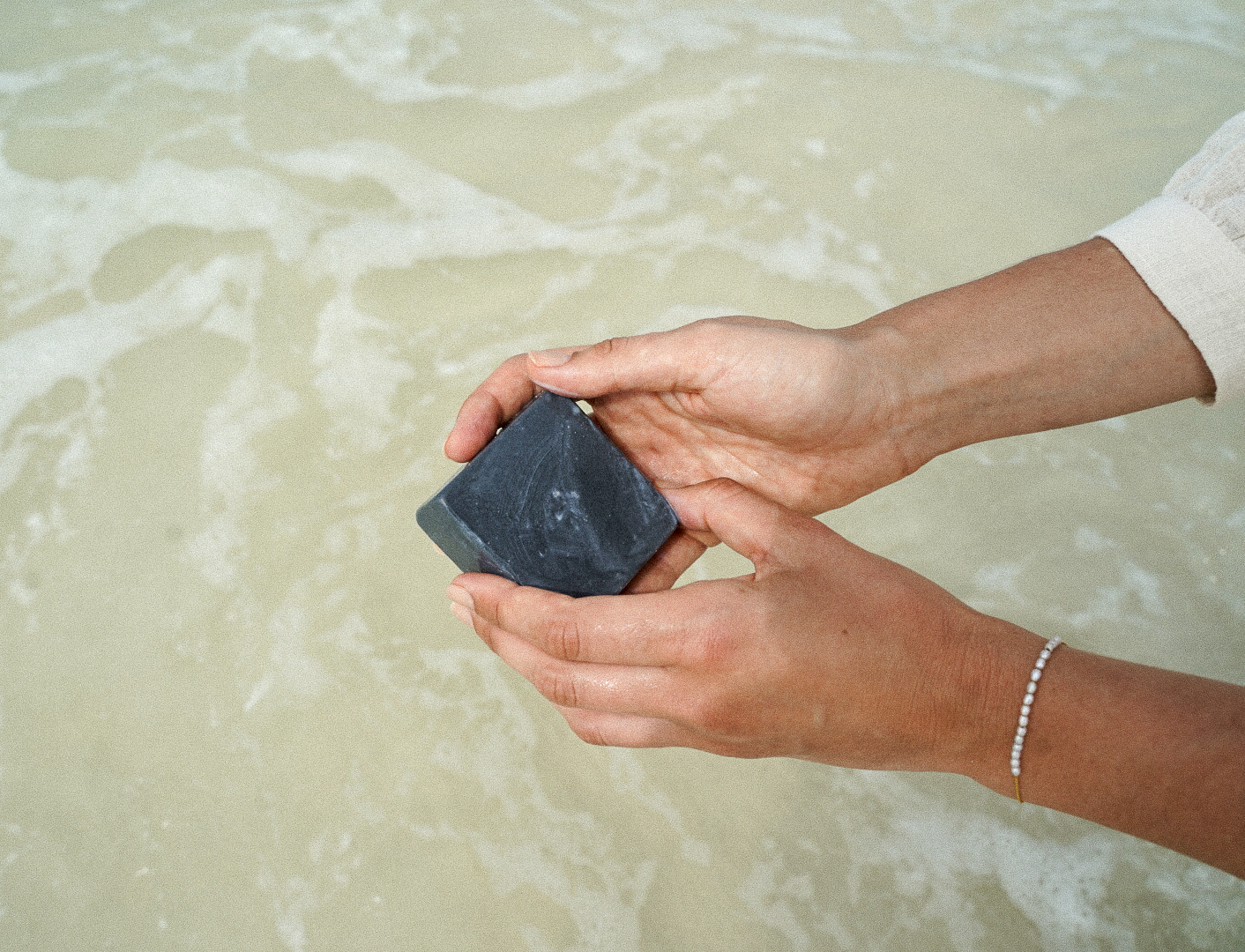 Hands hold black soap bar above shallow ocean water, sunlight glinting off skin for a serene, wellness-inspired beachside scene.