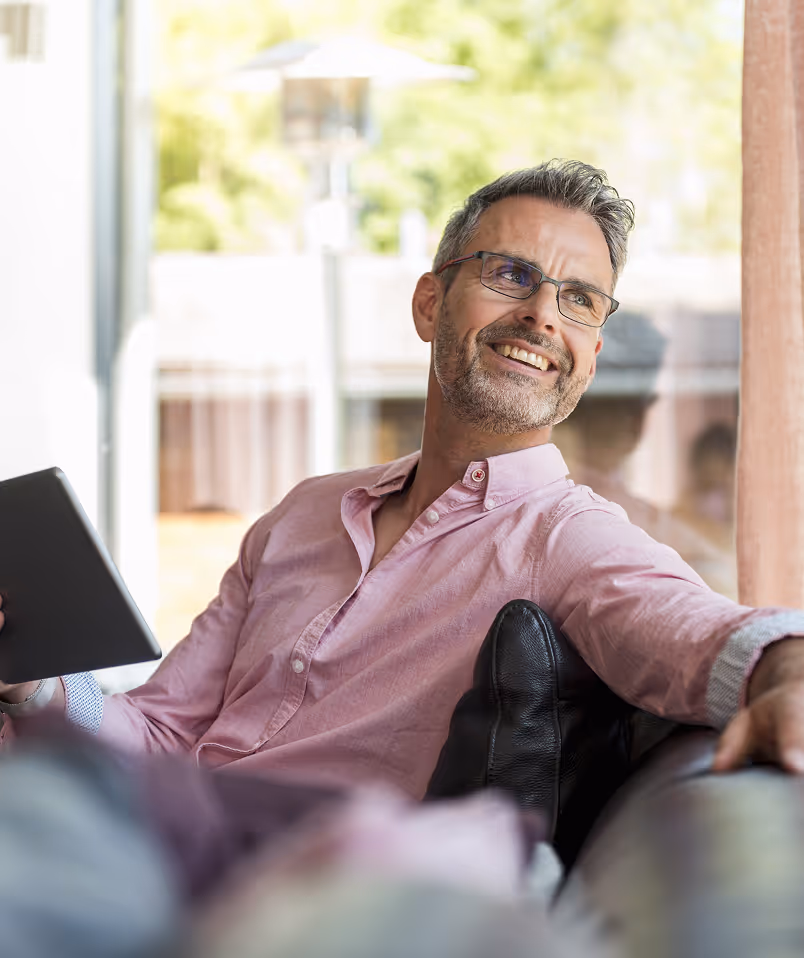Middle-aged man with salt-and-pepper hair and glasses, smiling while relaxing on a sofa and holding a tablet, with bright natural light and greenery in the background.