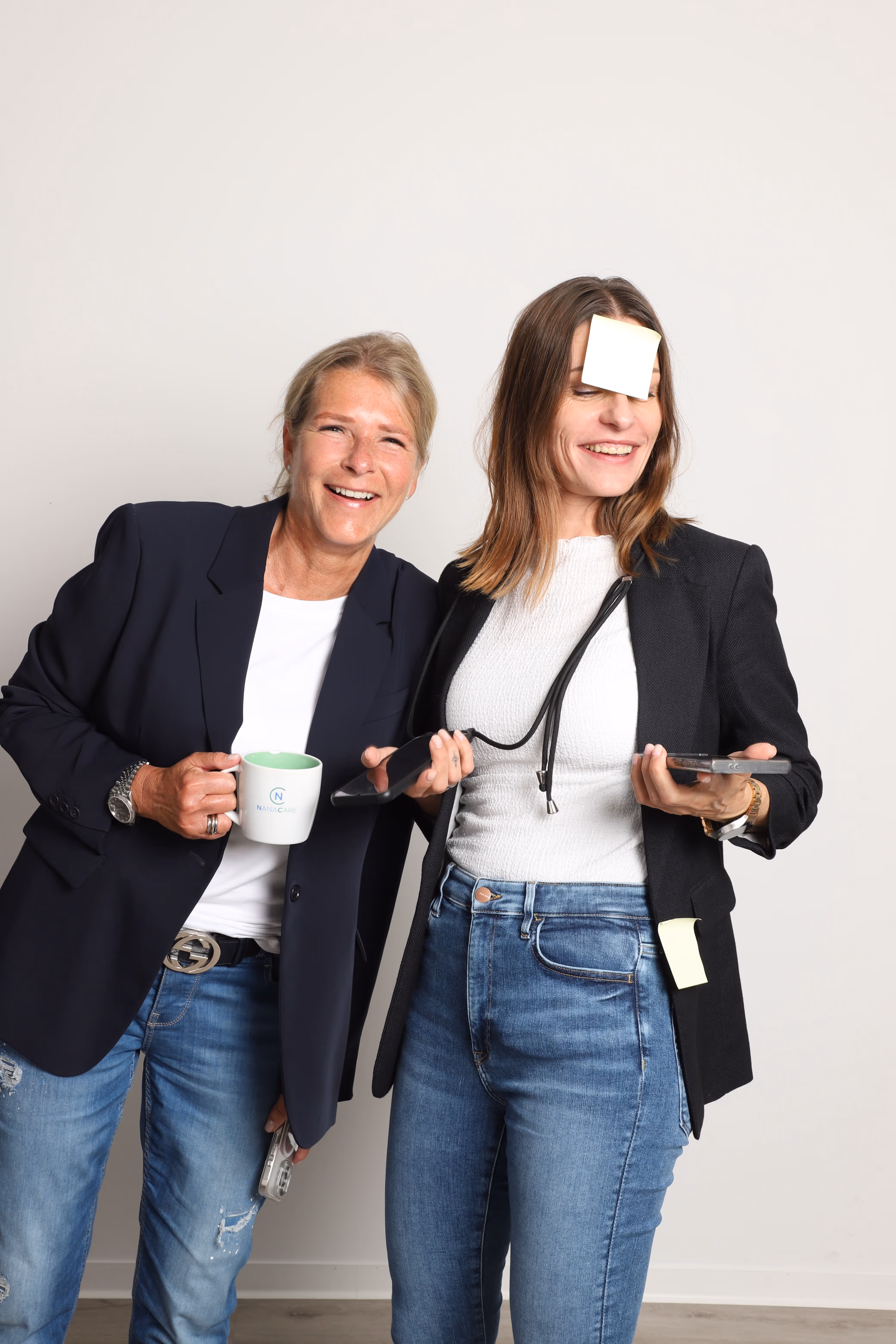 Die beiden Geschäftsführerinnen von Nana Care lächelnd vor hellem Hintergrund – eine mit Kaffeetasse, die andere mit Notizzetteln.“