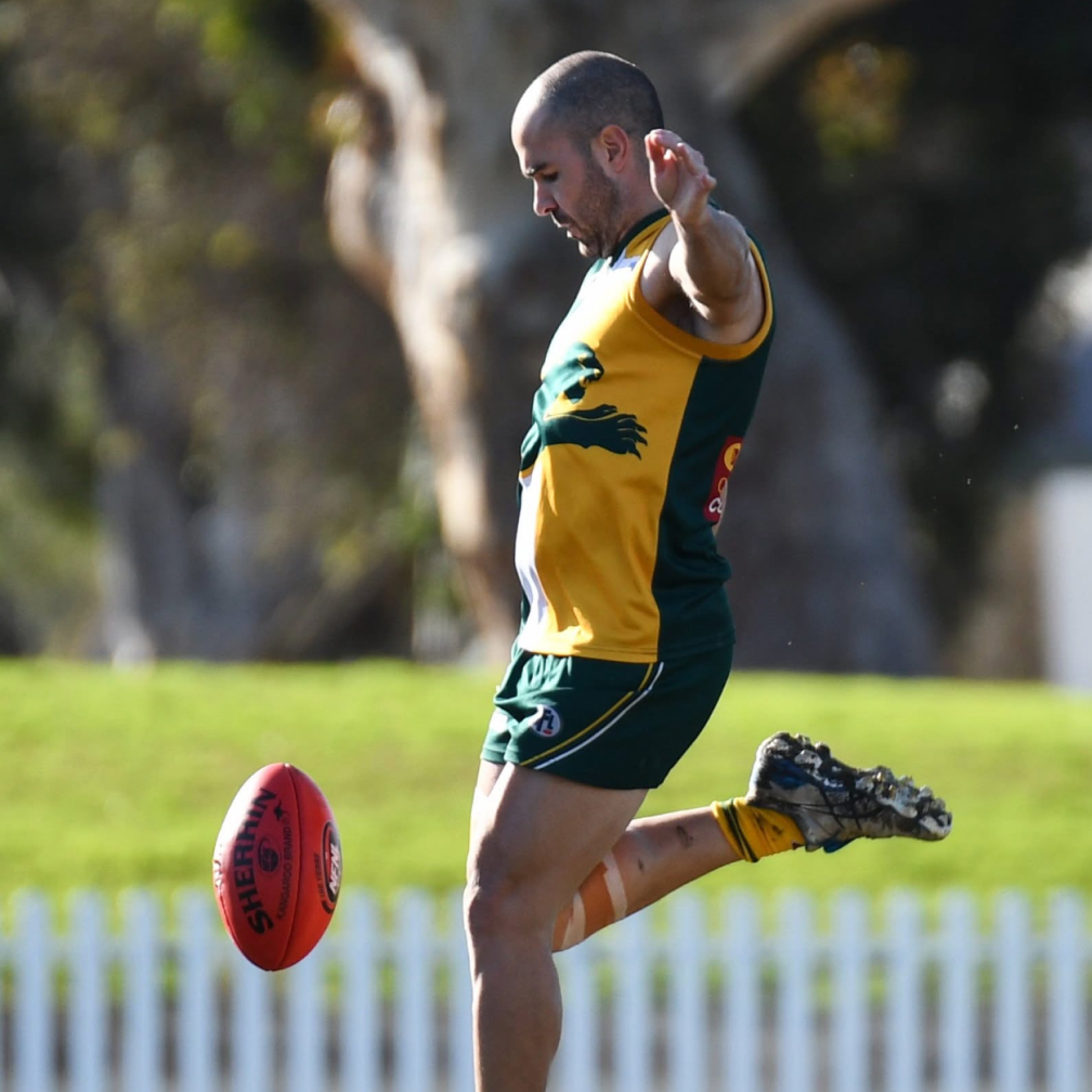 A male Australian rules football player in yellow and green uniform kicking a red football on a grassy field.