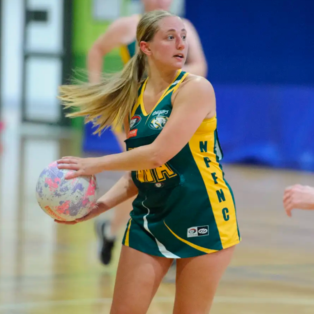 Female netball player in green and yellow uniform holding a ball while looking to her left on an indoor court.
