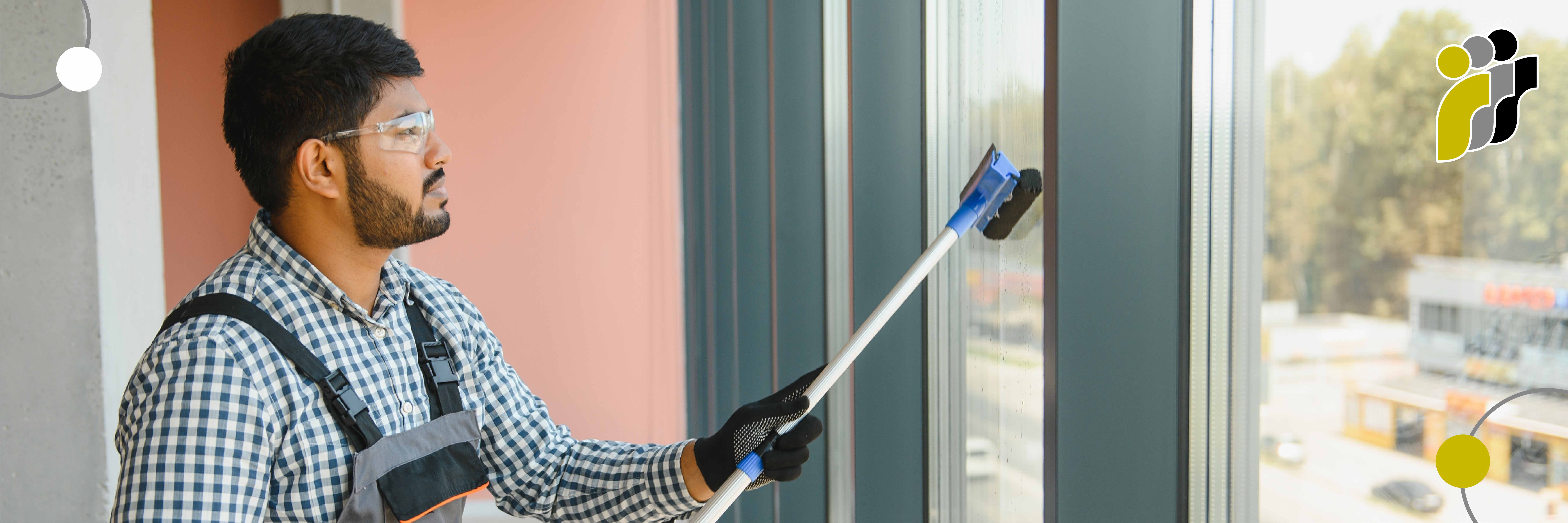 Ein Mann mit Schutzbrille und Handschuhen reinigt ein großes Fenster mit einem an einem langen Stiel befestigten Scheuerschwamm.