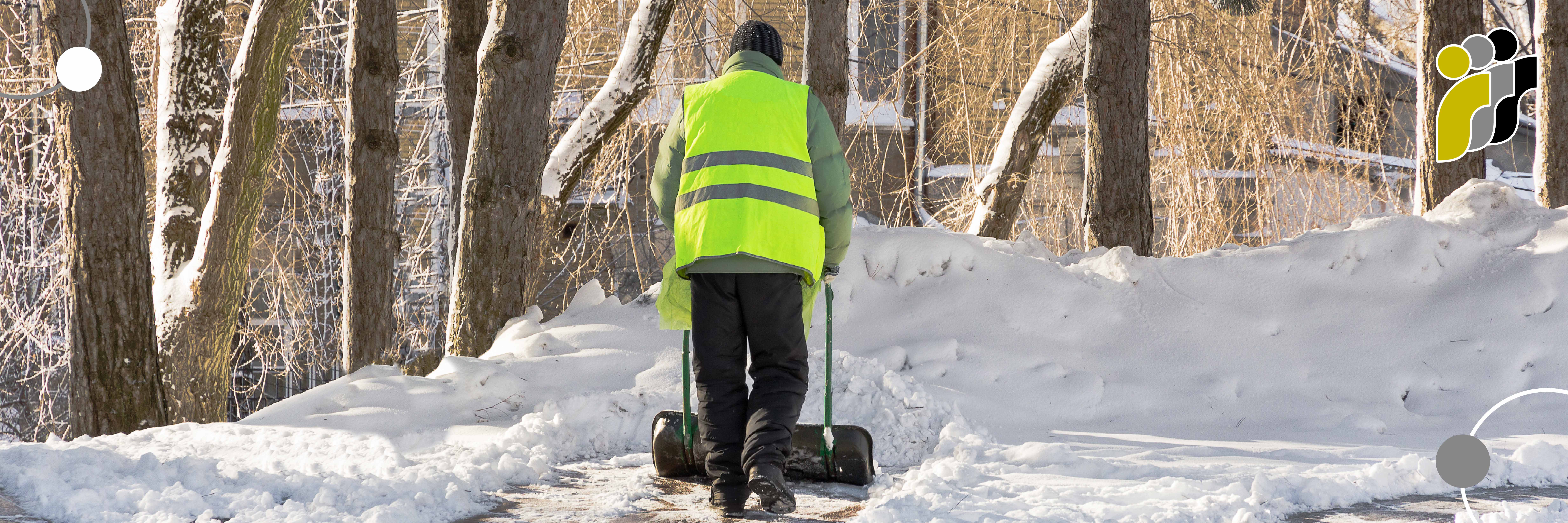 Eine Person in einer neongelben Sicherheitsweste schaufelt Schnee auf einem Weg; im Hintergrund sind schneebedeckte Bäume zu sehen.