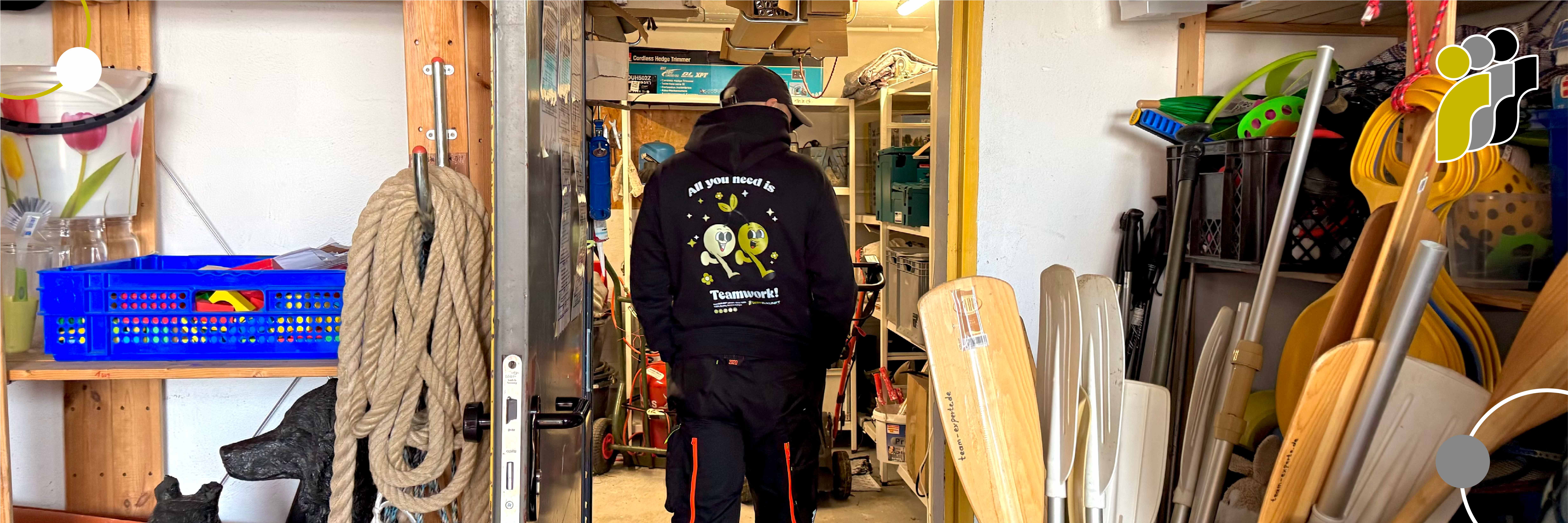 Eine Person, die einen schwarzen Kapuzenpullover mit der Aufschrift „All you need is Teamwork!“ auf dem Rücken trägt, steht in einem Lagerraum voller Seile, Paddel und verschiedener Ausrüstungsgegenstände.