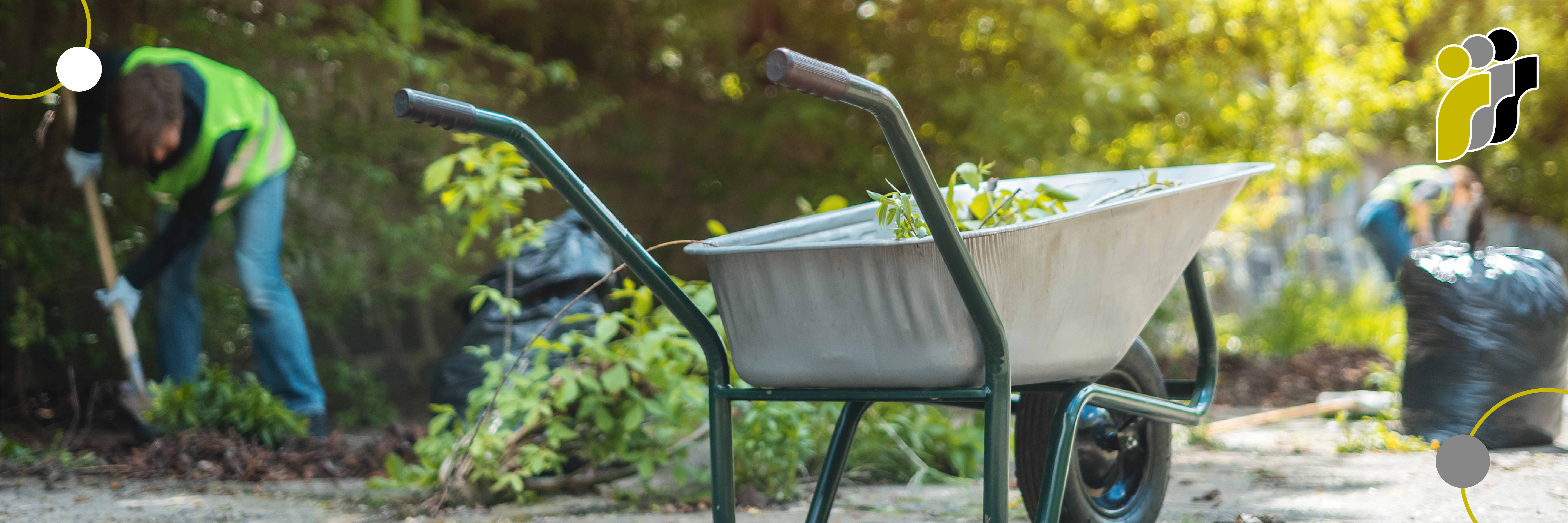 Eine Schubkarre voller Gartenabfälle; im Hintergrund arbeiten zwei Personen in Sicherheitswesten im Garten.