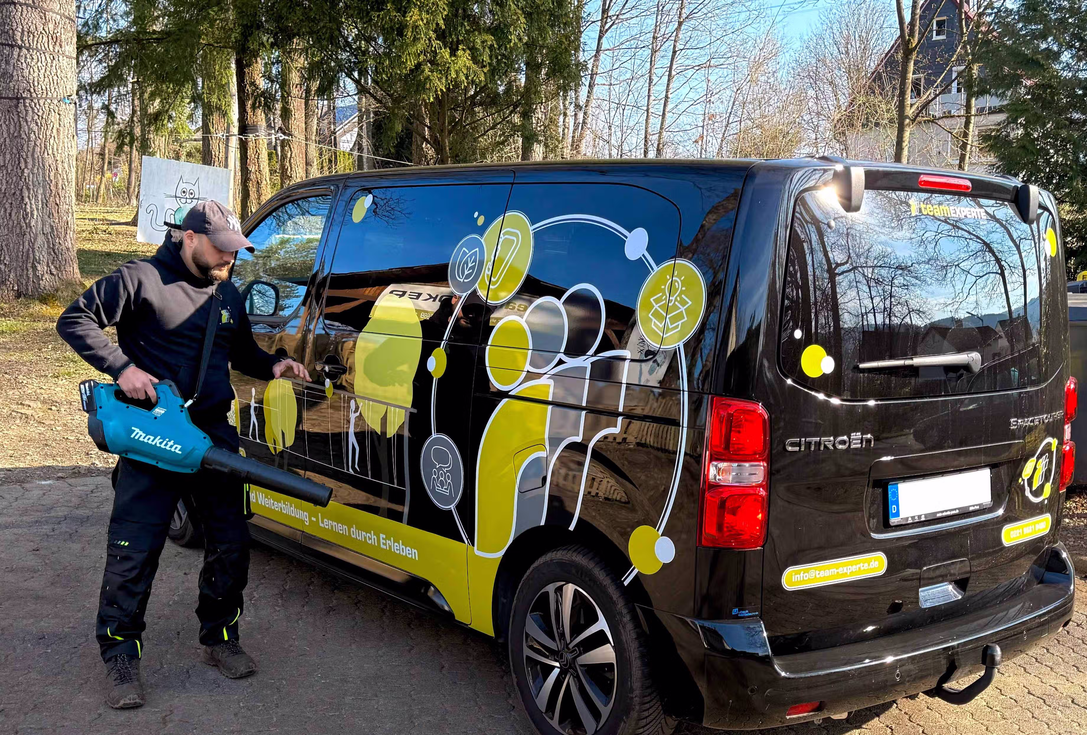 Ein Mann benutzt einen blauen Makita-Laubbläser neben einem schwarzen Citroën-Transporter mit gelb-weißer Aufschrift.