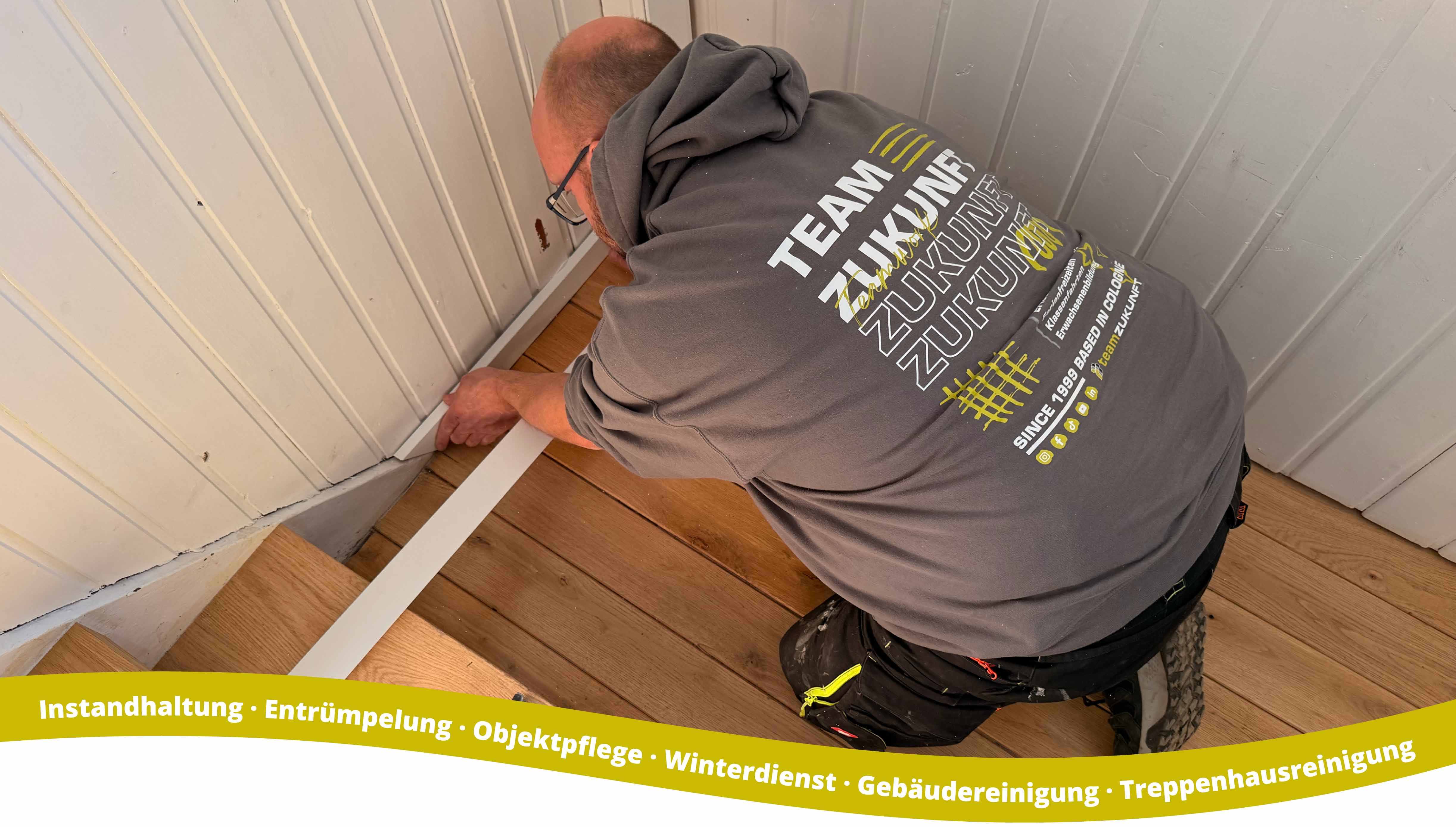 A man wearing a gray hoodie with the text 'TEAM ZUKUNFT' on the back is installing or adjusting white trim along the corner where a wooden floor meets a white paneled wall.