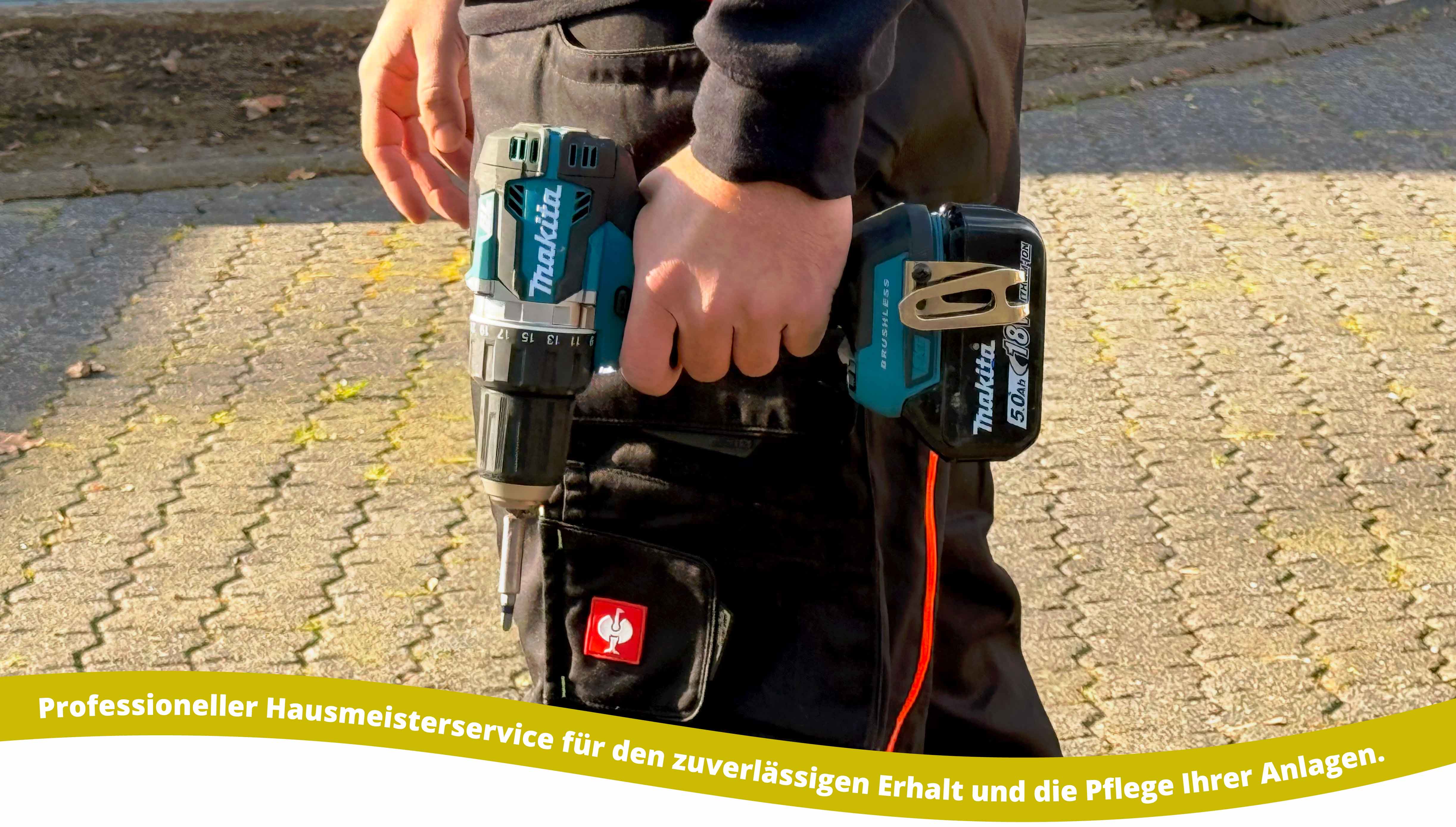 Person holding a Makita cordless drill outdoors, wearing work pants with a red tool logo on the pocket, standing on a paved surface.
