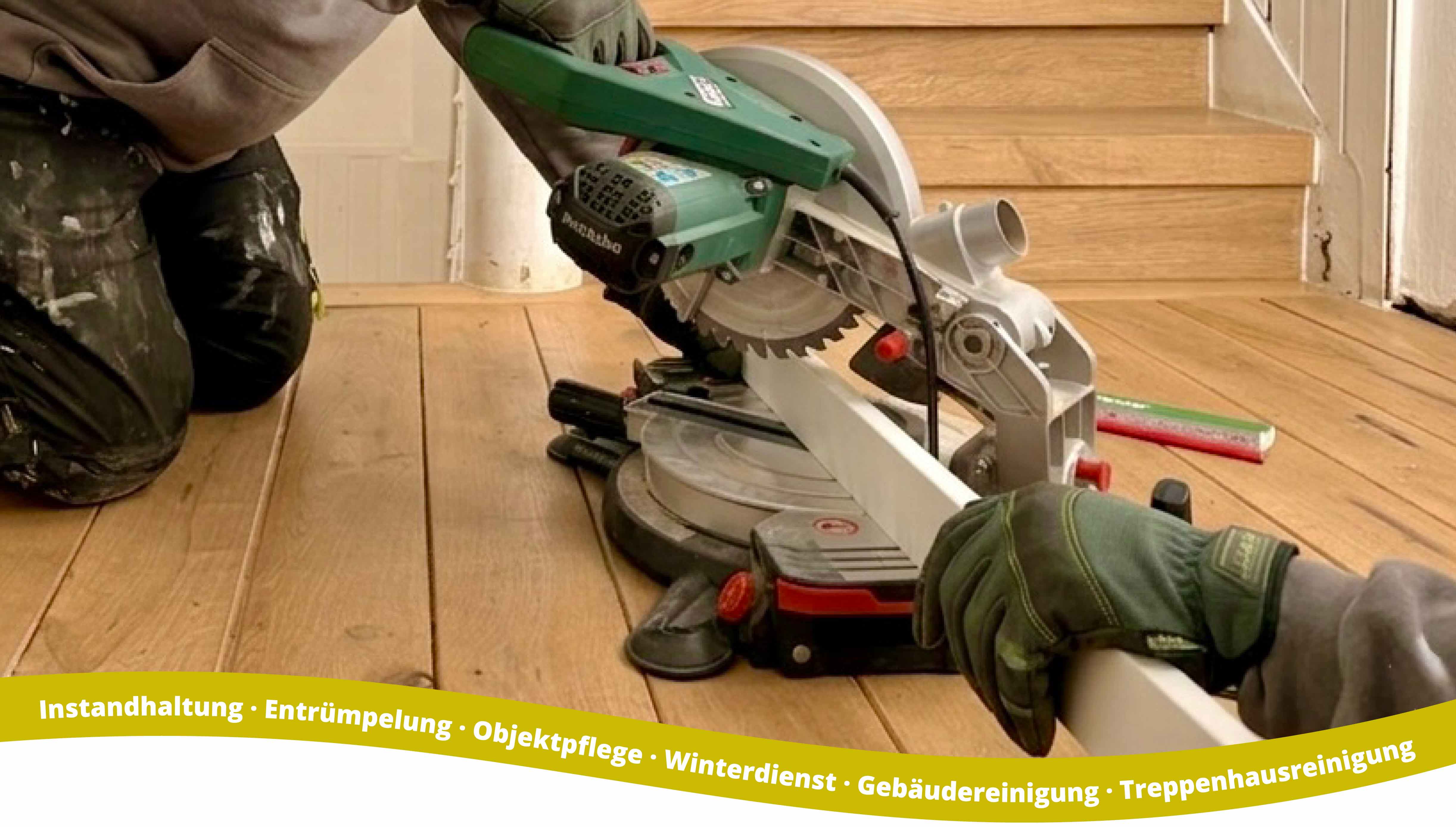 Person wearing gloves and work clothes using a green and white miter saw to cut a white wooden plank on a wooden floor near wooden stairs.