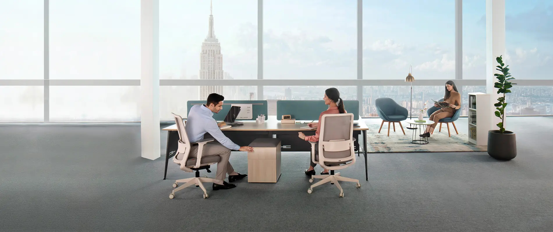 Office with two people sitting at desks and one person reading in a lounge area near large windows.