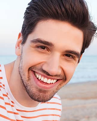 Junger Mann mit strahlend weißen Zähnen lächelt am Strand nach professioneller Zahnaufhellung bei den Q-Zahnärzten Freiburg.