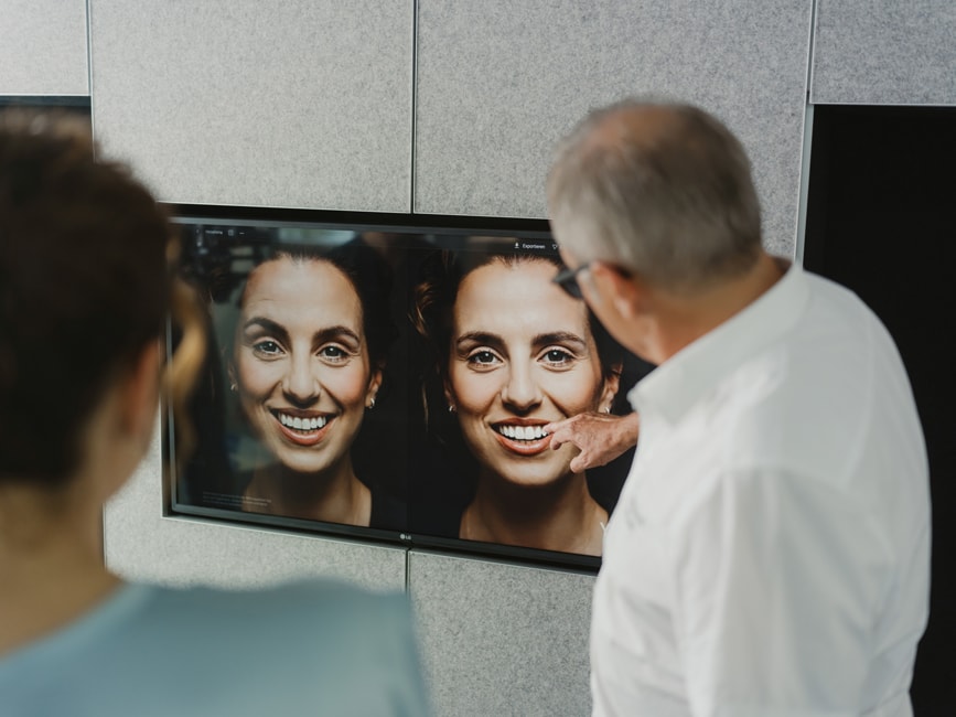 Veneers Freiburg – Zahnarzt Dr. Ralf Quirin zeigt Patientin den Vorher-Nachher-Vergleich des geplanten Veneers-Lächelns auf dem Bildschirm.