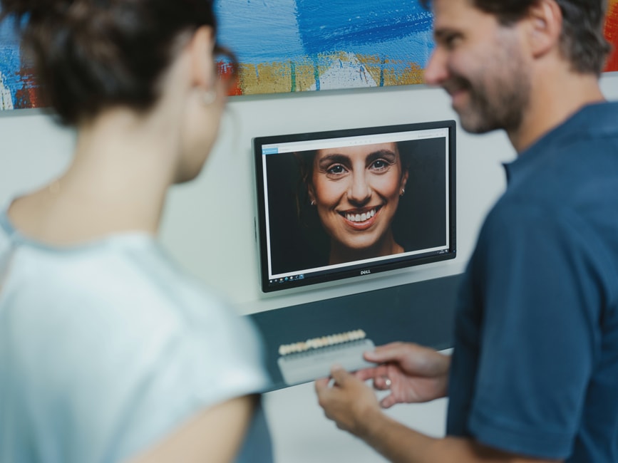 Professionelles Bleaching in Freiburg – Zahnarzt zeigt der Patientin am Monitor die geplante Zahnaufhellung und vergleicht die Ziel-Zahnfarbe mit einer Farbskala.