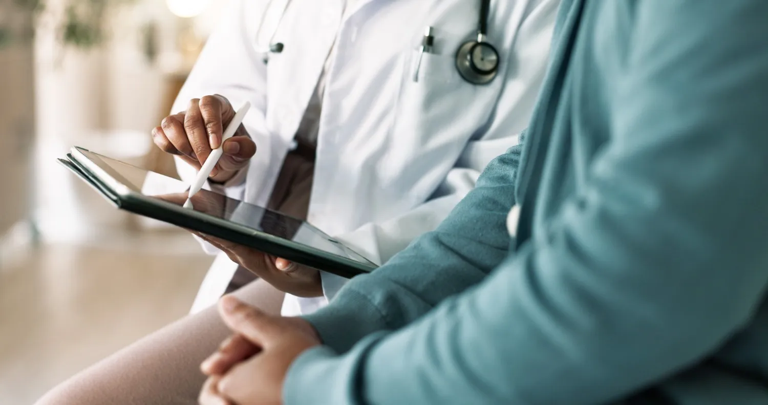 A doctor in a white coat with a stethoscope is showing a patient a medical chart on a tablet.
