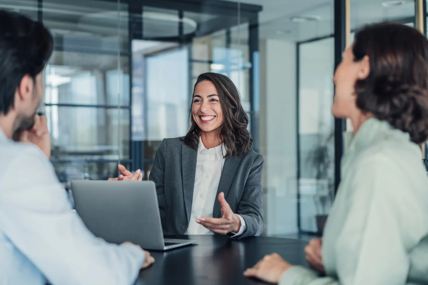A businesswoman is smiling and gesturing during a discussion with two colleagues at a table with a laptop.