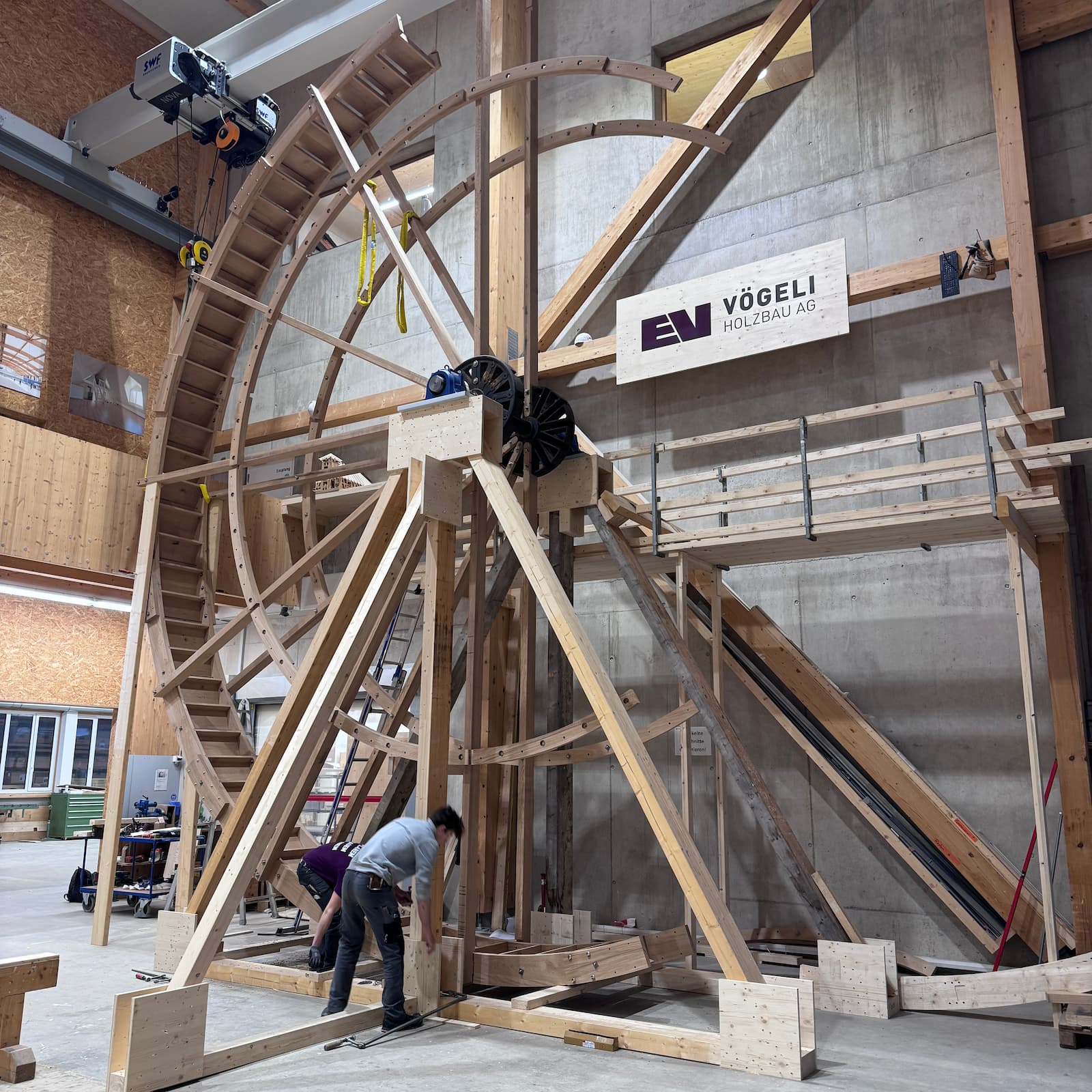 In der Produktionshalle der Vögeli Holzbau AG arbeiten zwei Handwerker an der Montage eines riesigen Wasserrades aus Holz, das einmal das grösste in Europa sein wird, mit sichtbaren gebogenen Trägern und einer komplexen Holzstruktur