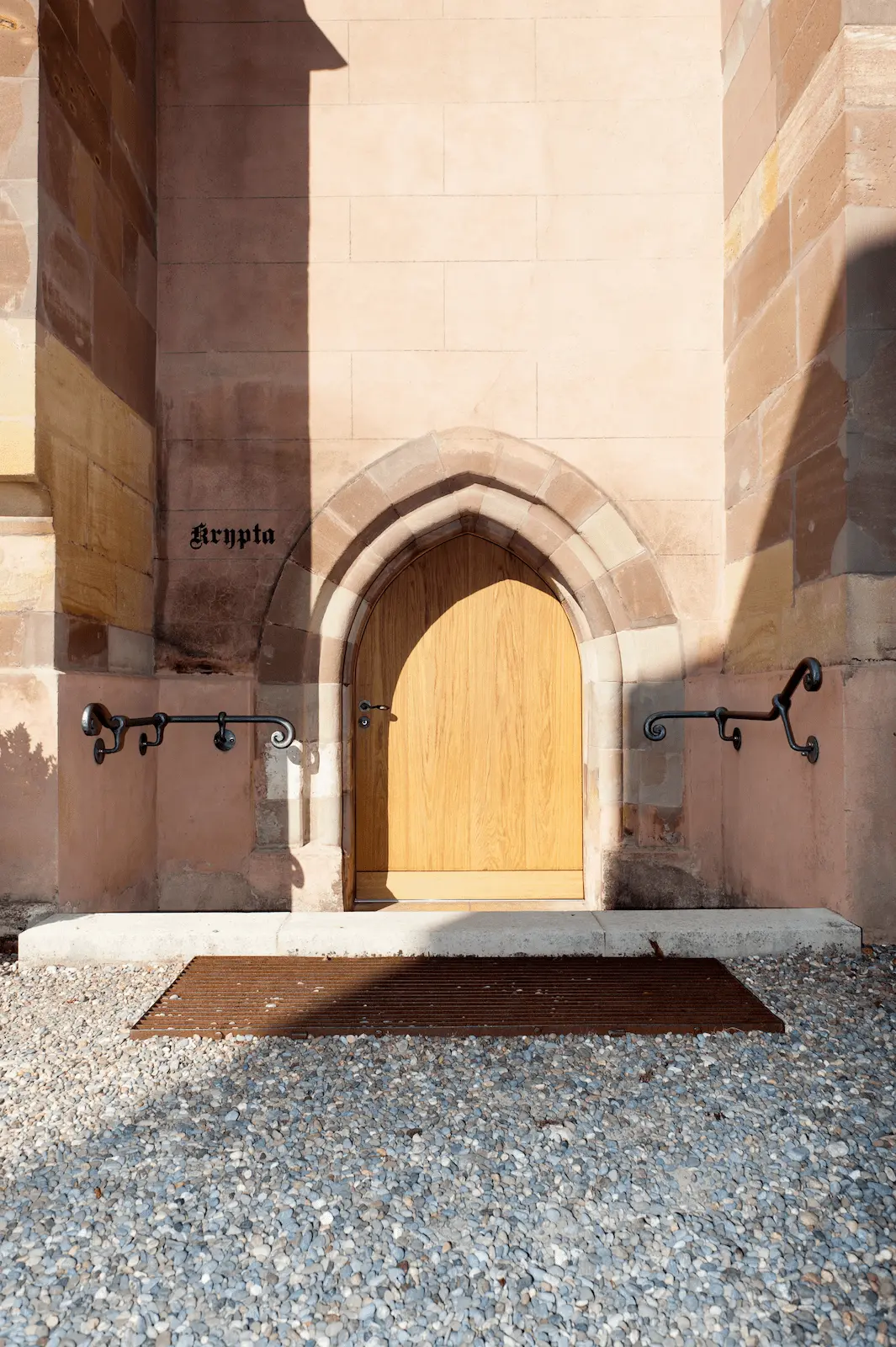 Gesamtansicht des Eingangs zur Krypta des Verenamünsters Bad Zurzach mit neuer Eichentüre, flankiert von historischen Steinmauern und dekorativen Geländern.