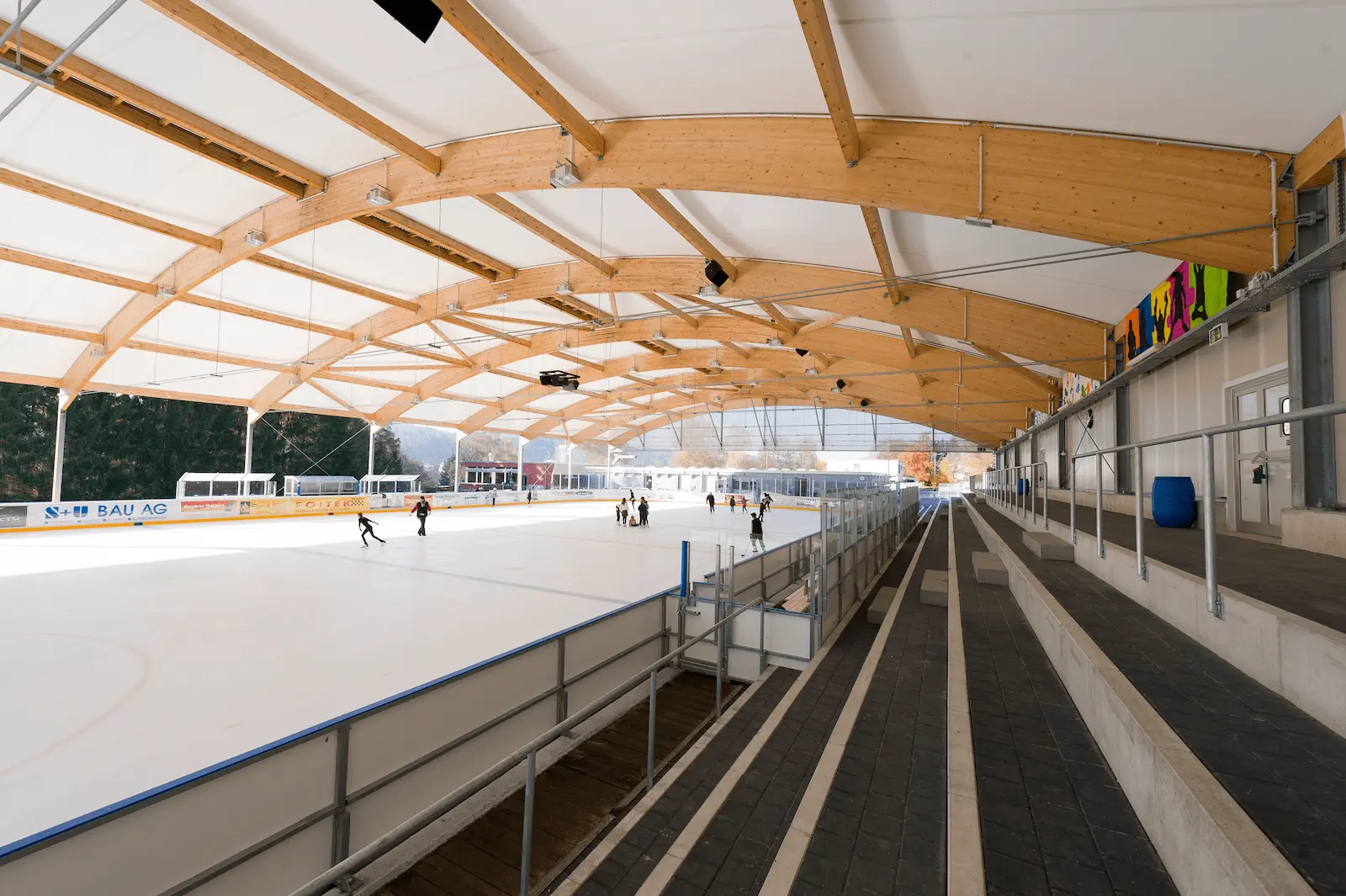 Innenansicht der Eisbahn Urdorf mit Holzbindern und Eisläufern auf der Fläche