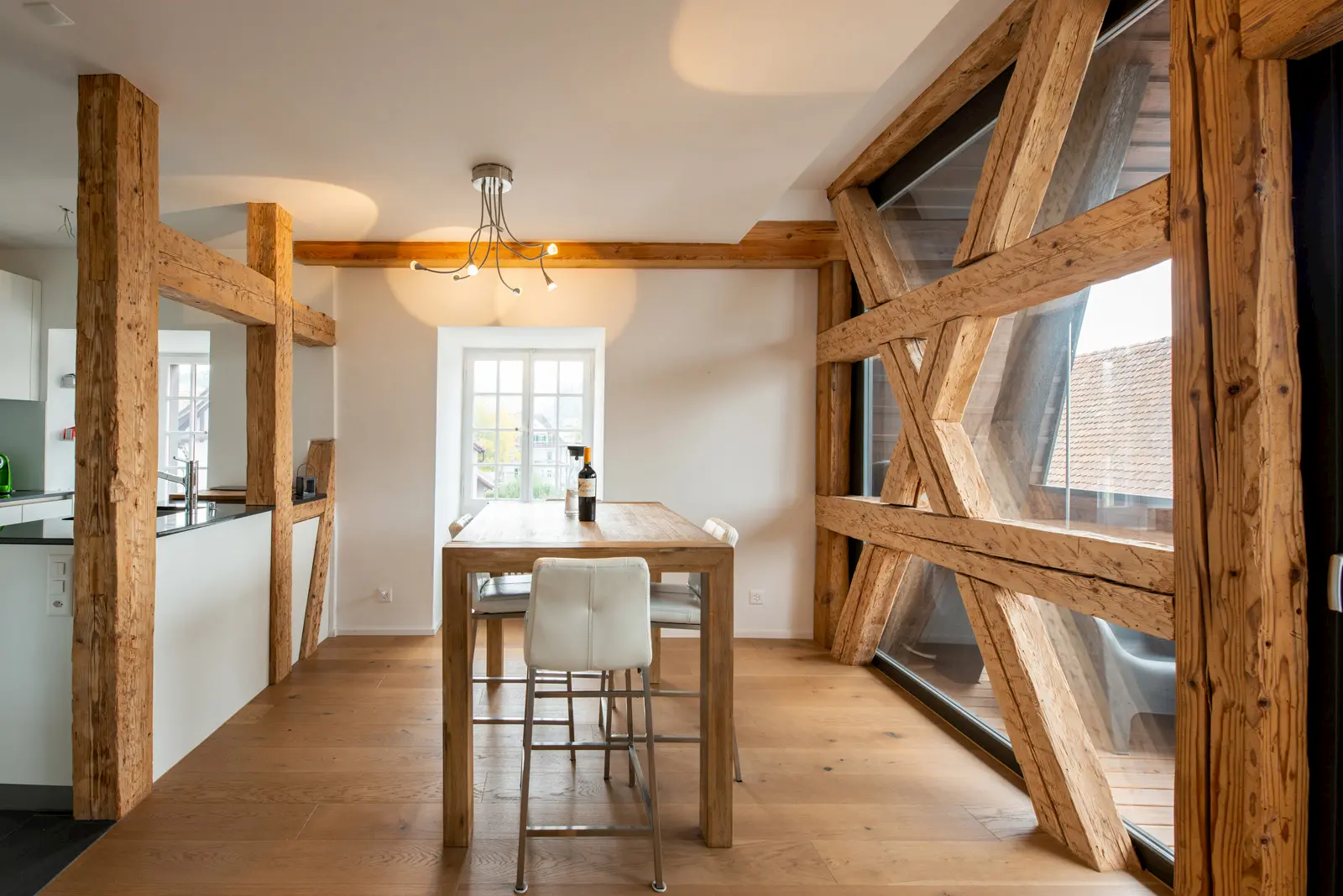 Heller Essbereich mit rustikalen Holzbalken, modernem Tisch aus Massivholz und großer Fensterfront mit sichtbarer historischer Holzkonstruktion.