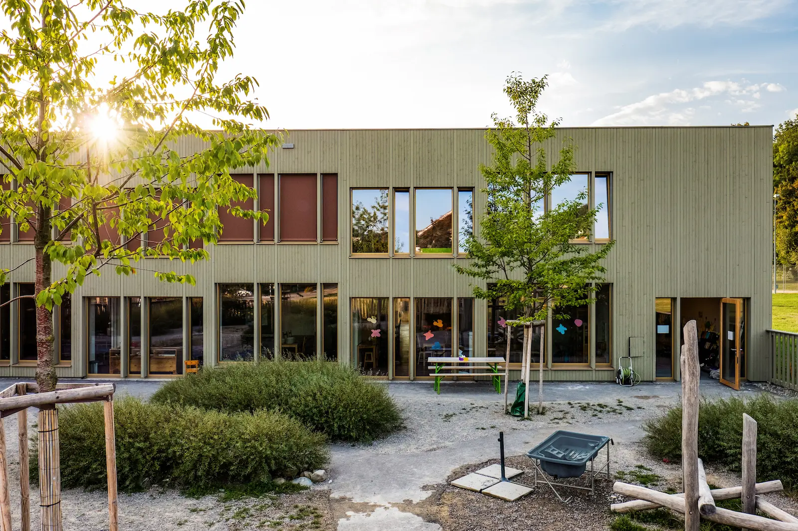 Rückansicht des Chinderhuus Goldiland mit stehender Holzfassade in Nut- und Kamm-Verbindung. Die Fassade ist mit Keim-Farbe behandelt und bietet optimalen Witterungsschutz. Im Vordergrund ist der Gartenbereich mit Bäumen, Sträuchern und Spielbereich für Kinder zu sehen, was die Verbindung von Architektur und Natur unterstreicht