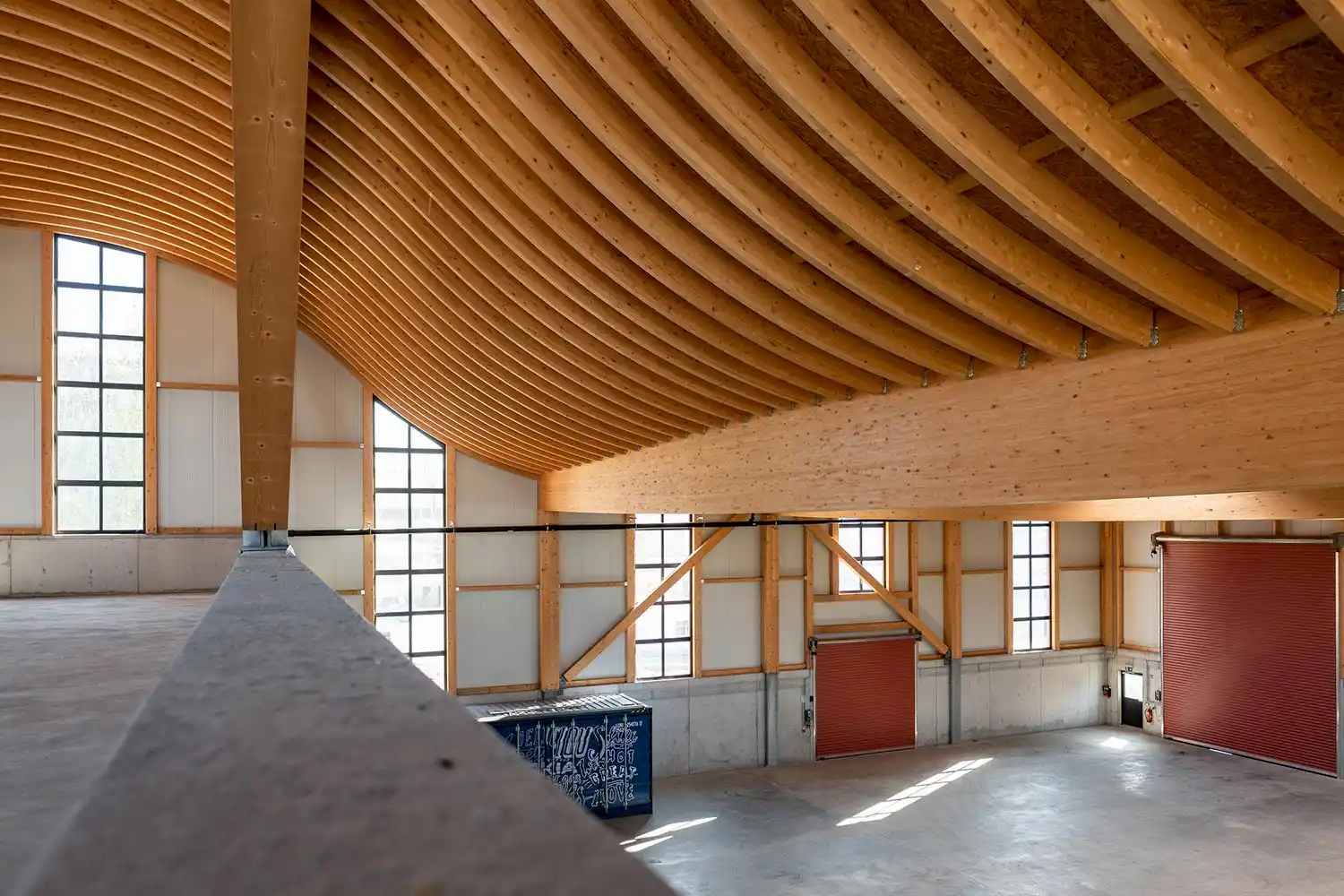 Innenansicht einer Halle mit geschwungenen Holztragwerken, großen Fensterfronten und sichtbarer Holzkonstruktion.