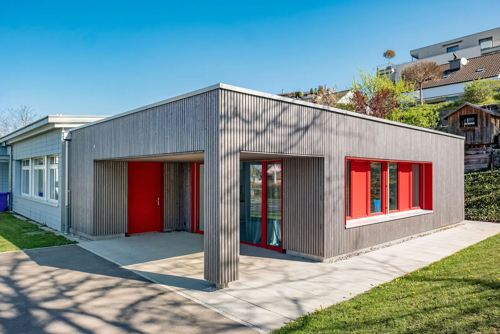Erweiterungsbau Kindergarten Clemenz mit Holzfassade und roten Fensterrahmen.