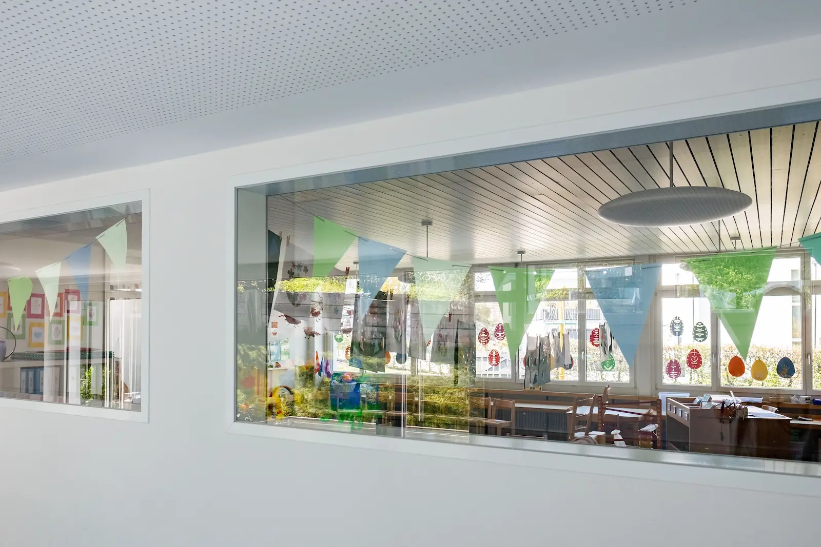 Blick durch Innenfenster in dekorierten Gruppenraum mit bunten Girlanden.