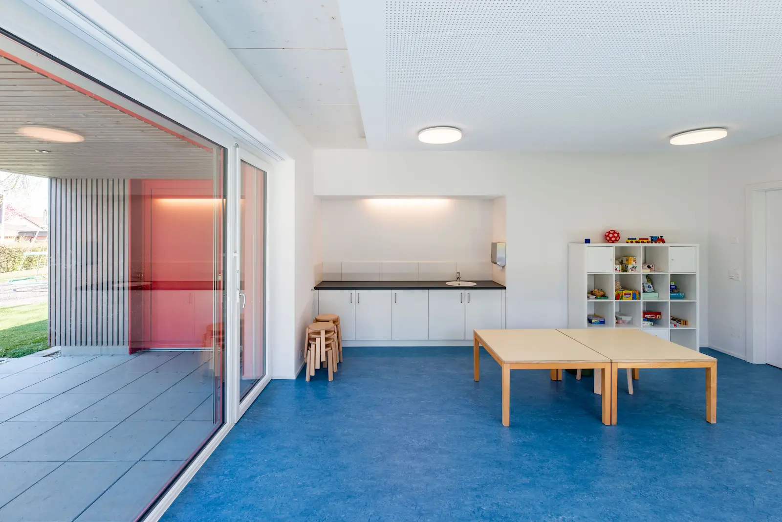 Innenraum Kindergarten mit blauem Boden, Holztischen und Regalwand.