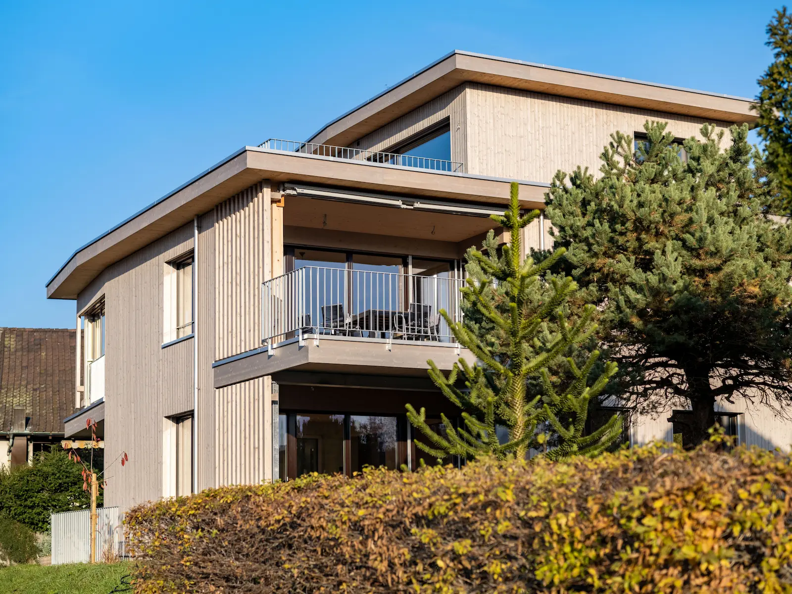 Seitenansicht eines Einfamilienhauses mit Holzfassade in hellem Grauton, Flachdach und grossen Fensteröffnungen.