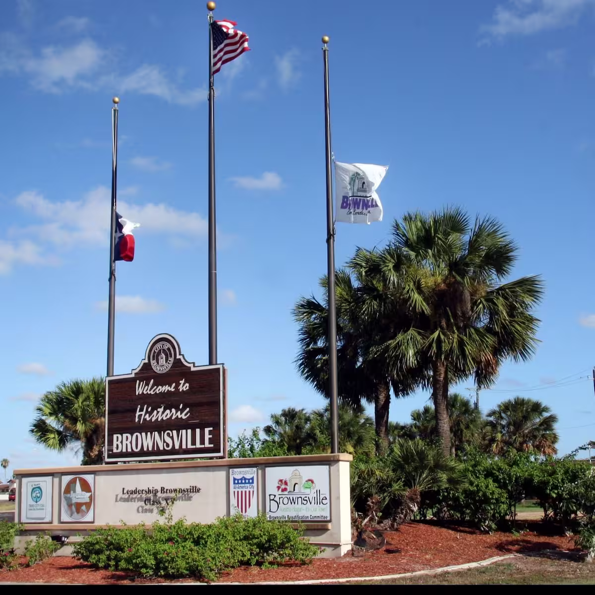 Welcome sign for Historic Brownsville with American, Texas, and city flags on flagpoles under a blue sky and palm trees.