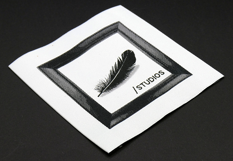 White woven label with a black feather and the /STUDIOS logo