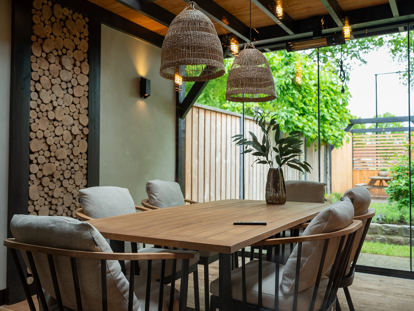 Sfeervolle overkapping met houten eettafel, rotan lampen en natuurlijke elementen, ingericht als buitenruimte voor overleg of ontspanning.